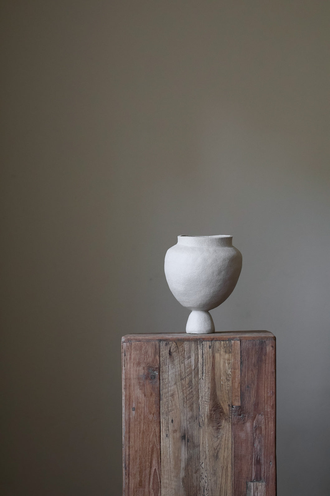 A hand-built, sculptural vase by Hana, crafted from a light-colored clay, leans slightly to the right.  It features a wide, almost spherical body that rests upon a narrow, pedestal-like base. The surface of the vase appears to have a slightly textured, matte finish, indicative of the hand-building process